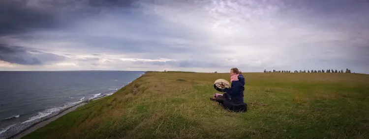 Verena sitzt mit Rahmentrommel auf einer Wiese an der Steilküste, im Hintergrund eine Steinsetzung